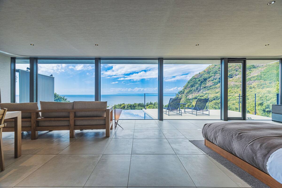 Ein geräumiges Schlafzimmer mit einem großen Bett, einem Holztisch und einem Stuhl. Es verfügt über ein großes Fenster mit Blick auf eine gebirgige Landschaft und ein Gewässer.