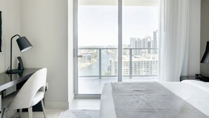 Une chambre d’hôtel moderne dotée d’un grand balcon, d’un bureau avec une lampe, d’un lit et offrant une vue sur le paysage urbain.