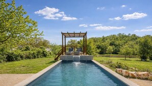Una piscina rectangular con una pérgola de madera y dos sillas de descanso.