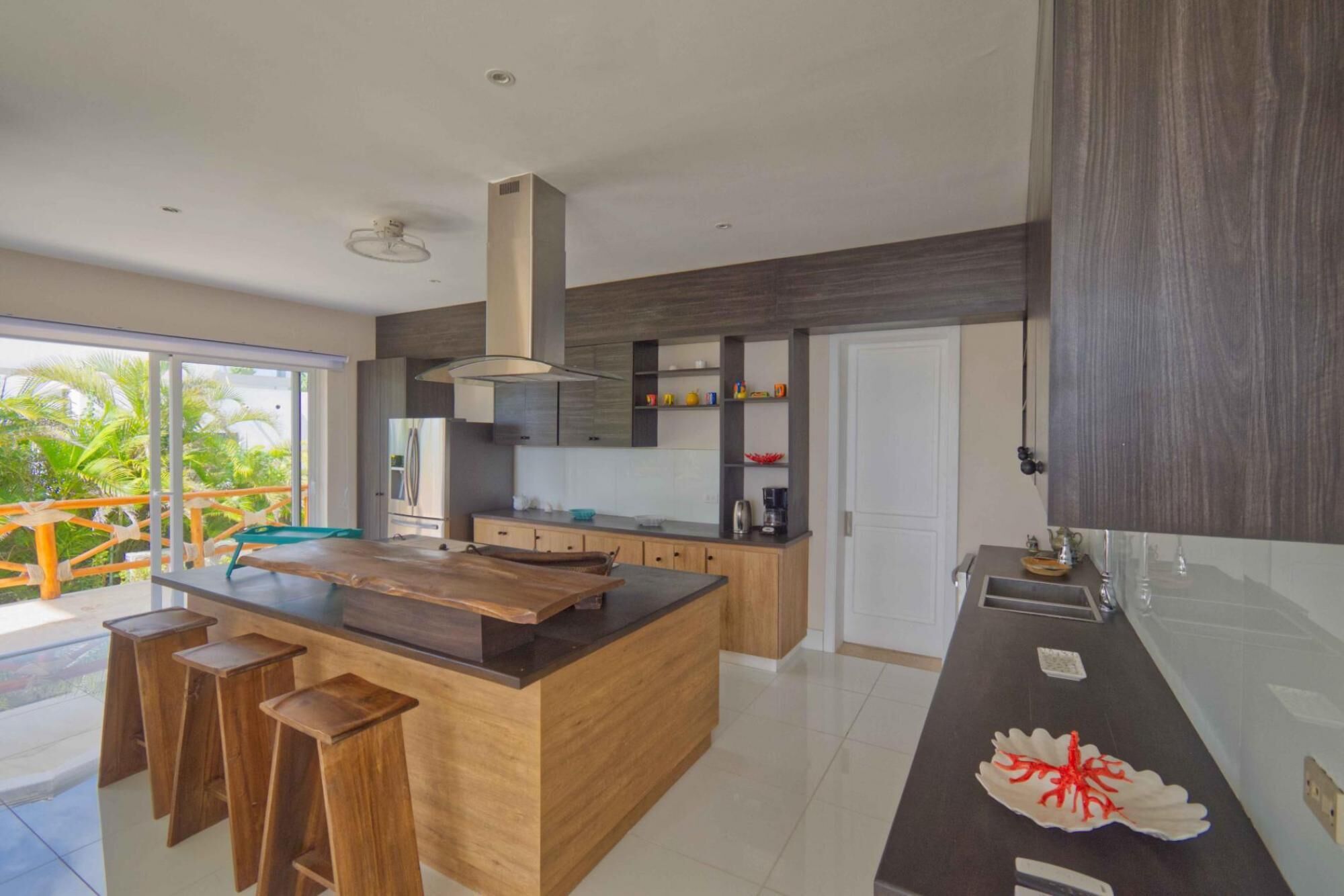 A modern kitchen with a central island, wooden stools, and a large window overlooking greenery.