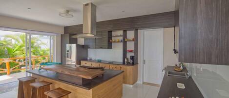 A modern kitchen with a central island, wooden stools, and a large window overlooking greenery.