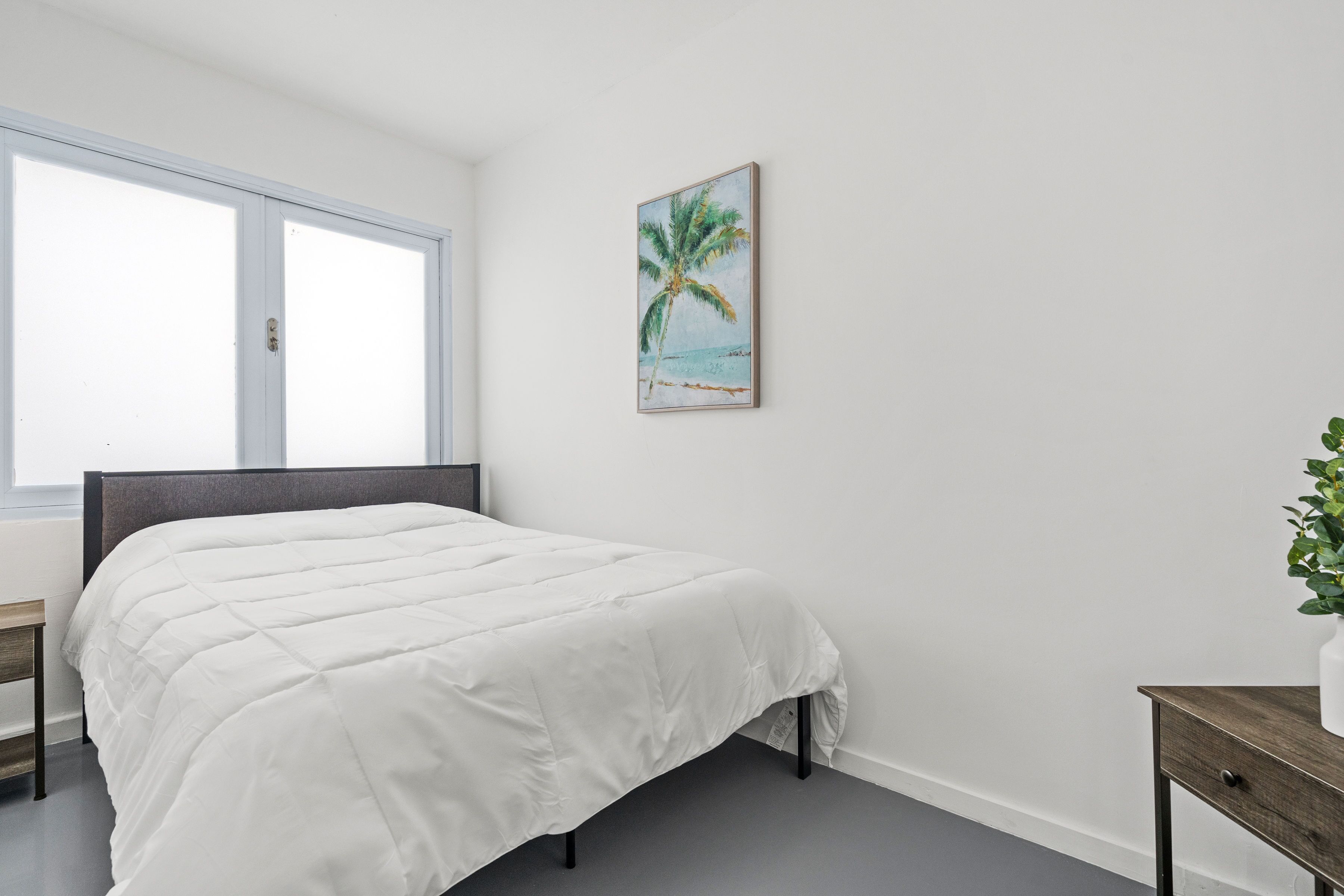 A bedroom with a bed, a nightstand, a bedside table, a window, and a wall-mounted painting of a palm tree.