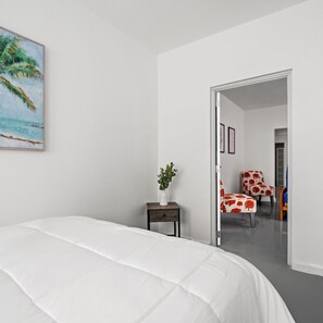 Une chambre à coucher comprenant un lit, une table de chevet, une plante en pot et une porte donnant sur une autre pièce où se trouvent une chaise et une table.