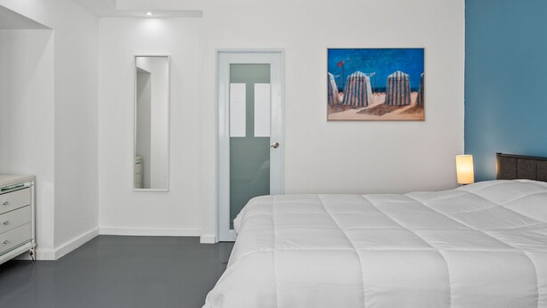 A bedroom with a large bed, a door, a mirror, a painting, and a dresser.
