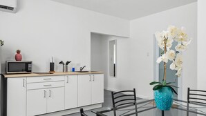 A modern kitchen with white cabinets, a microwave, and a sink. There is a dining table with chairs and a vase of white orchids.