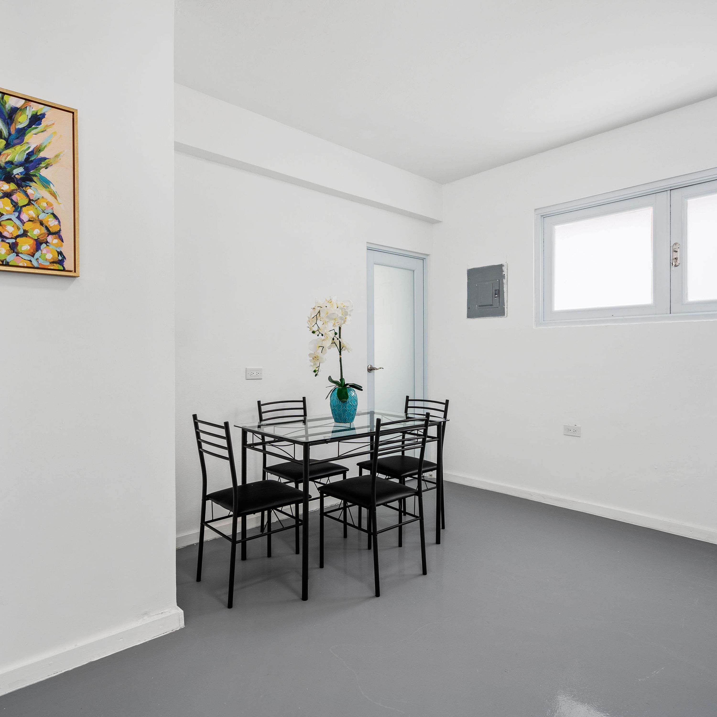 A dining area with a glass table and four chairs, a vase with flowers, and a painting on the wall.
