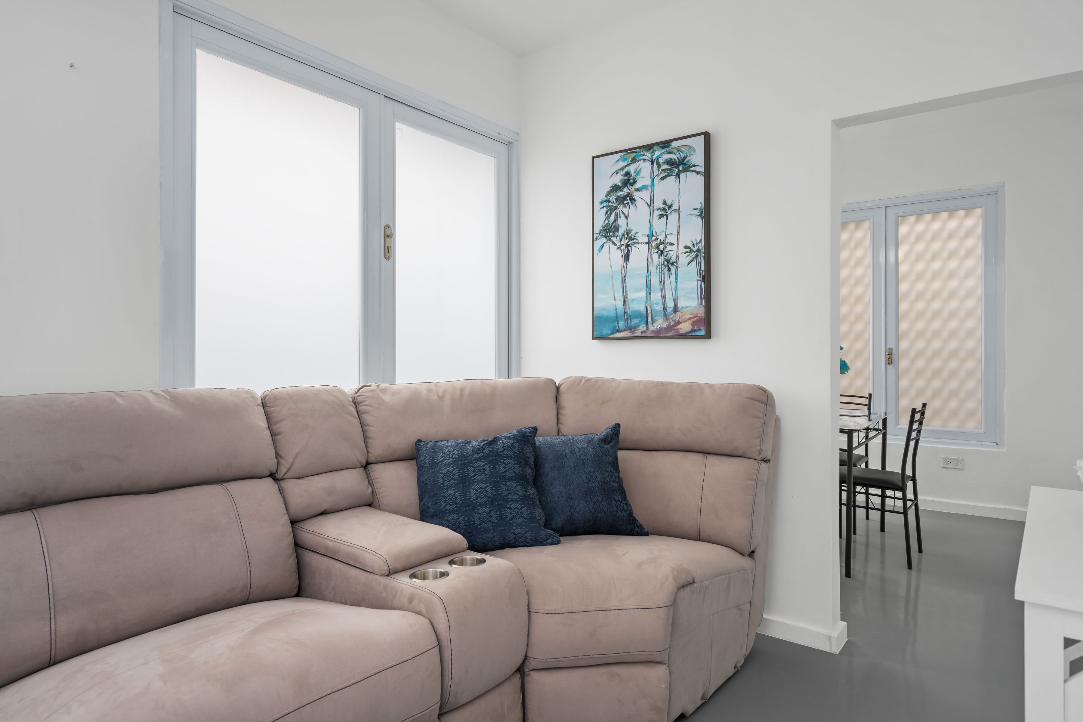 A beige sectional sofa with two dark blue pillows, a dining table with chairs, and a painting of palm trees on the wall.