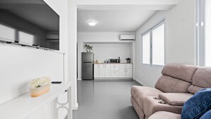 A modern living room with a sofa, a TV on a white shelf, and a kitchen area with a refrigerator and microwave. - Otium Rentals Best Condado Location Near Beach (San Juan)