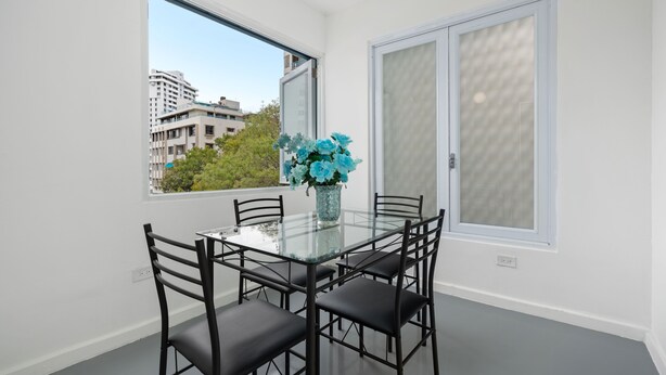 A dining area with a glass table and black chairs, a vase with blue flowers, and a view of buildings and trees through large windows. - Otium Rentals Best Condado Location Near Beach (San Juan)