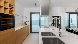 A modern kitchen with a large island, stainless steel sinks, and a view of the sea through large windows.