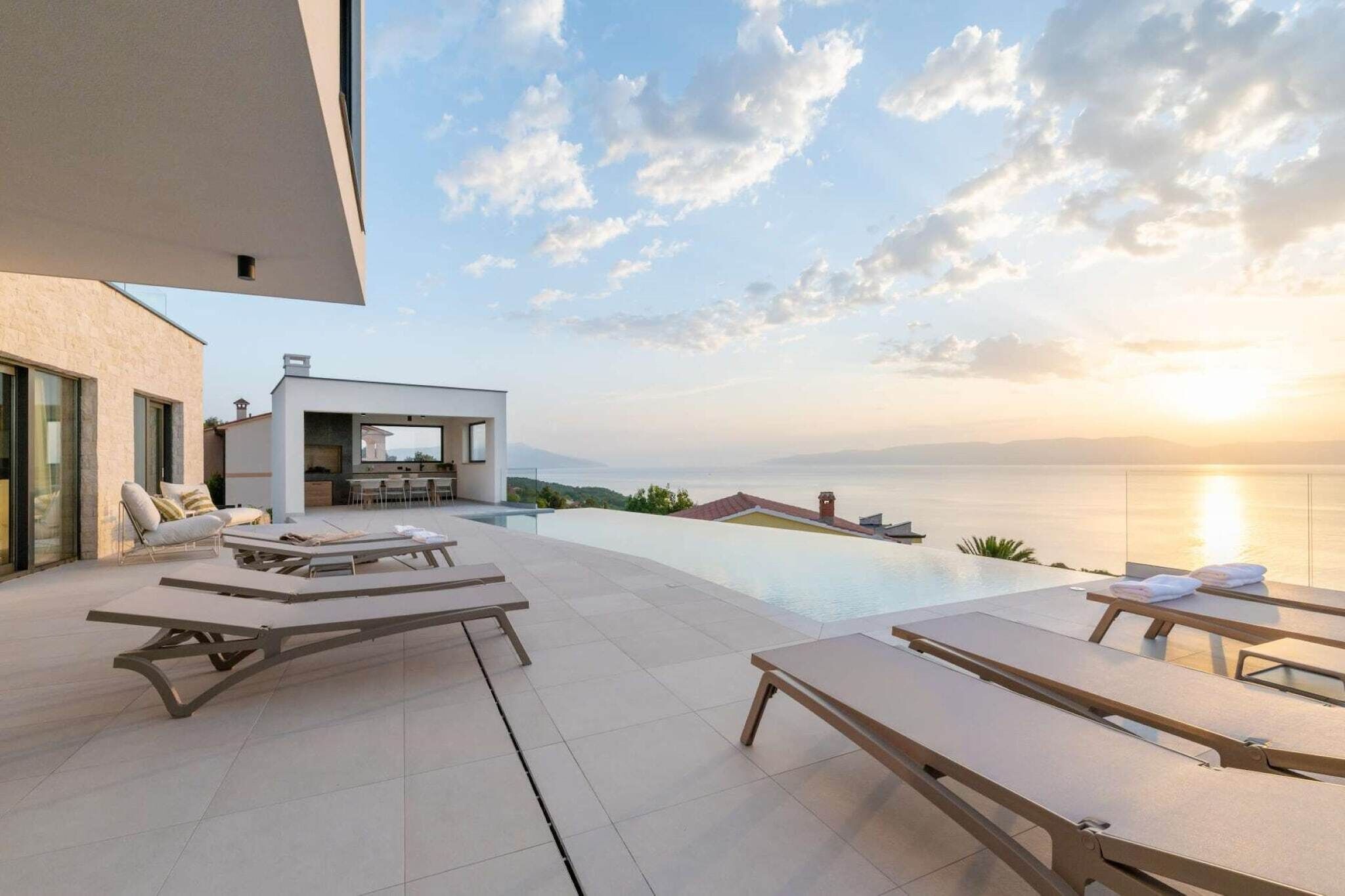 Un espace piscine extérieur moderne, doté de chaises longues, d’une cuisine et offrant une vue sur la mer au coucher du soleil.