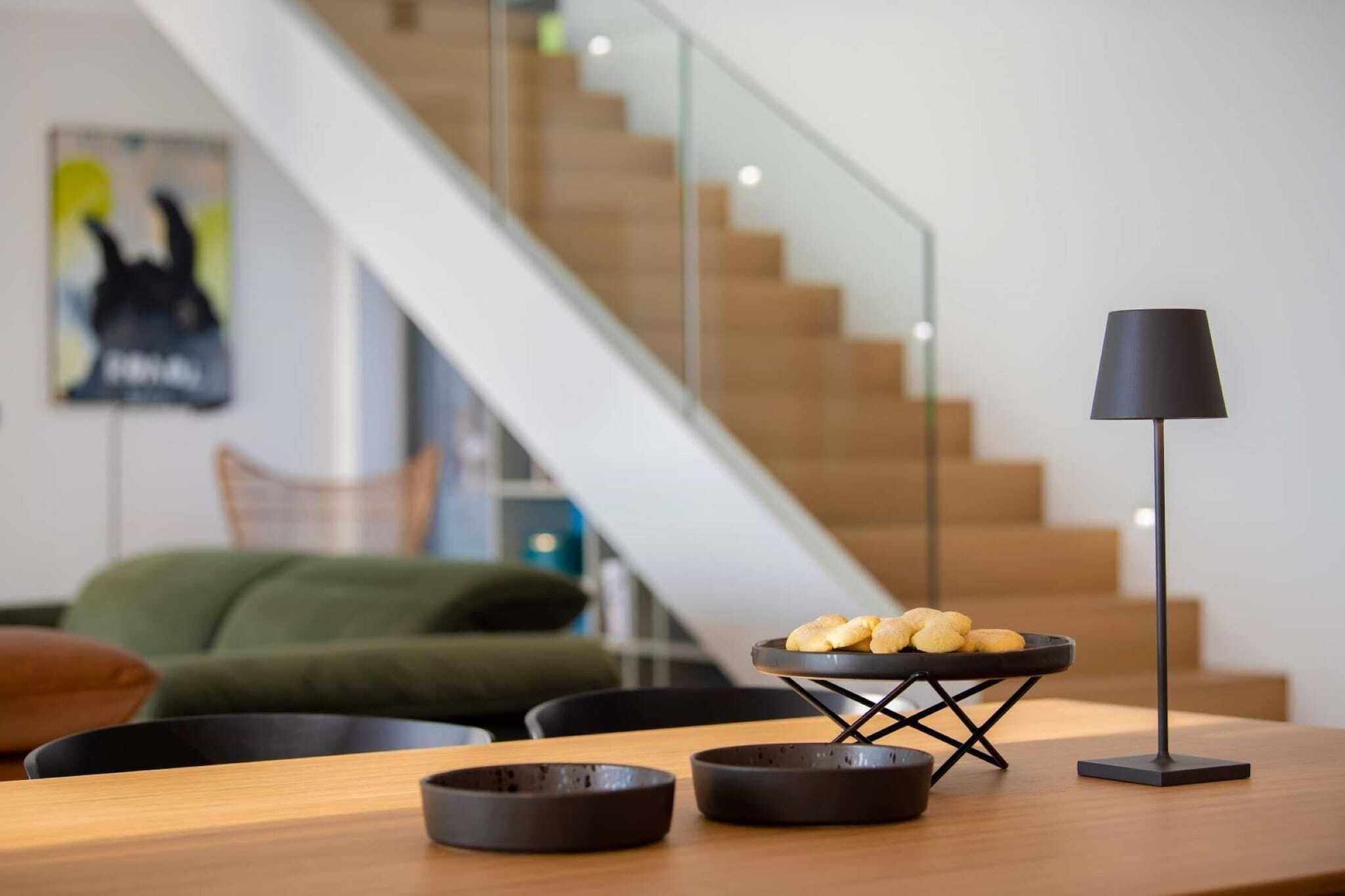 En moderne stue med et træbord, en sort lampe og en skål med snacks. En trappe er synlig i baggrunden.