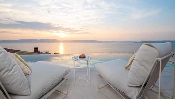 A serene poolside lounge with two white cushioned chairs, a small round table with a bowl and cups, and a view of the sunset over a calm body of water.