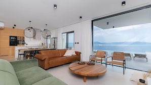 A modern living room with a large sofa, a round coffee table, and wicker chairs. There is a kitchen area with a dining table and a large window overlooking the sea.