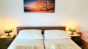 Una cama doble con dos almohadas blancas, una mesita de noche con una lámpara y un cuadro colgado en la pared de un árbol.