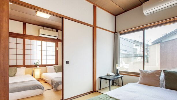 Ein traditionelles japanisches Hotelzimmer mit Tatami-Böden, einem verschiebbaren Shoji-Bildschirm, einem Bett mit grüner Bettdecke und einem kleinen Tisch mit Lampe.