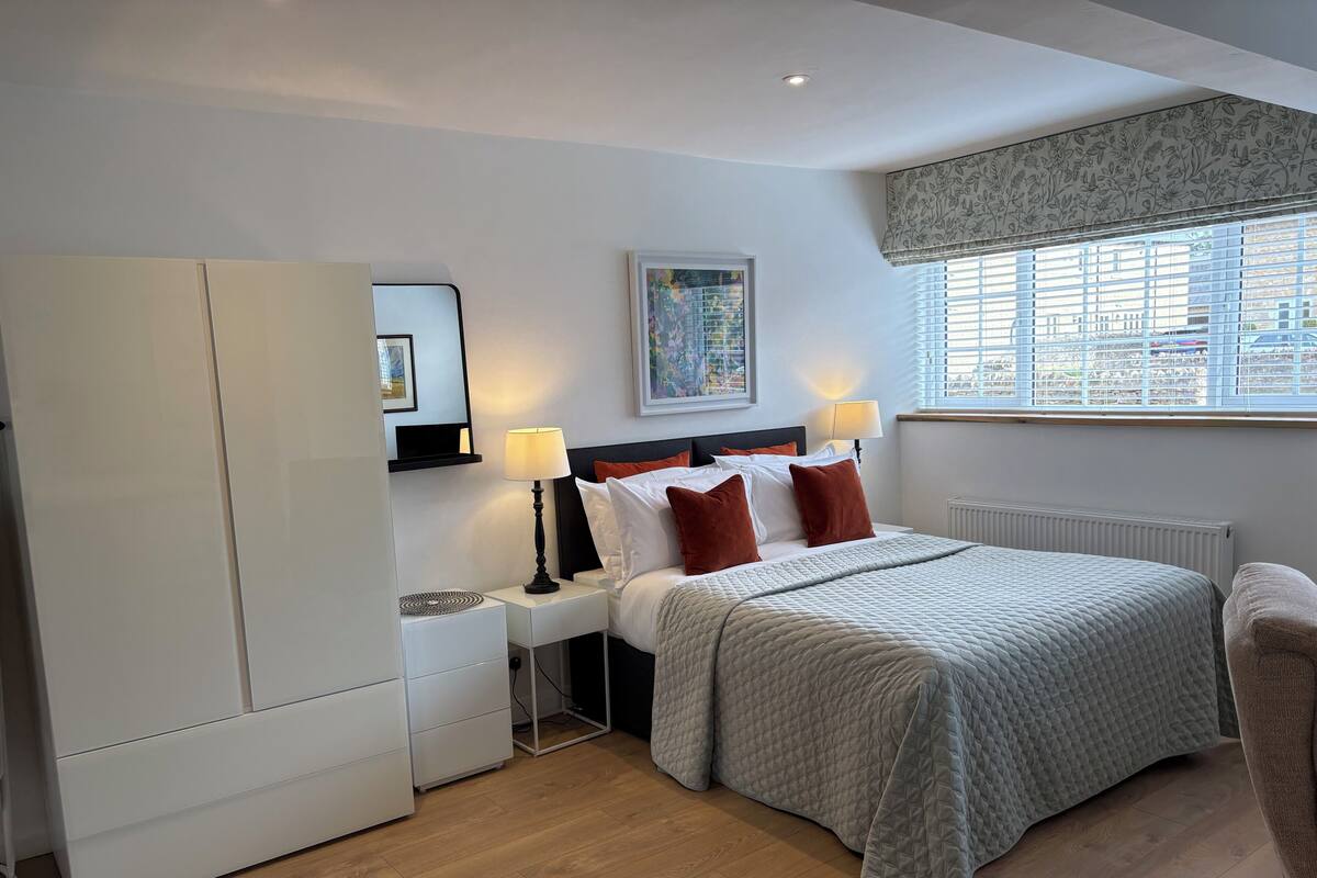 A hotel room with a bed, bedside tables, a wardrobe, a window with blinds, and a painting on the wall.