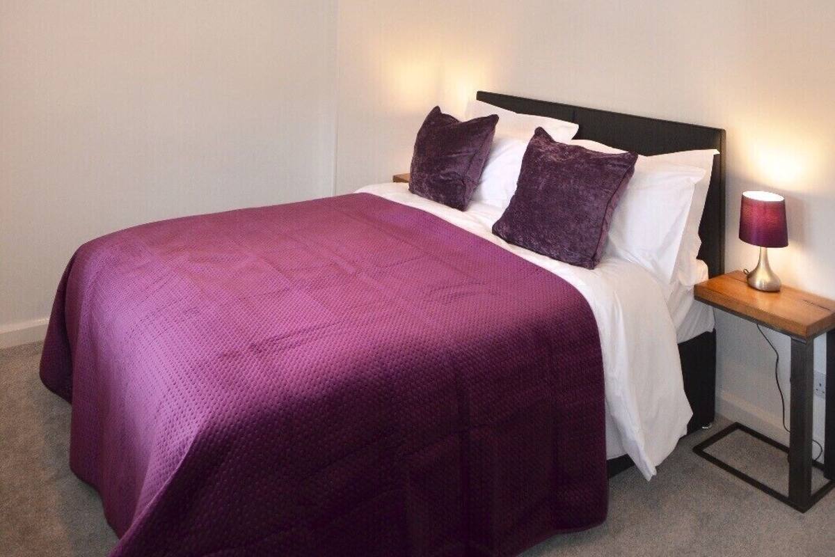 A bedroom with a bed, two purple pillows, a wooden nightstand with a lamp, and two chandeliers.