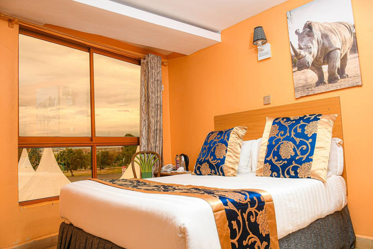 A hotel room with a large bed, a view of the sky through the window, a mounted picture of a rhinoceros, and a lamp on the wall.