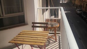 A balcony with a wooden table and chairs, a view of a street with parked cars, and a building in the background.