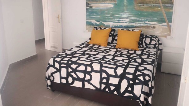 A bedroom with a bed featuring a black and white abstract pattern, two mustard-colored pillows, and a large tropical beach painting on the wall.