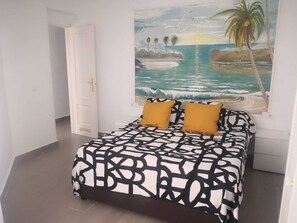 Ein Schlafzimmer mit einem Bett, das ein schwarz-weißes abstraktes Muster, zwei senfgelbe Kissen und ein großes Gemälde von einem tropischen Strand zeigt.