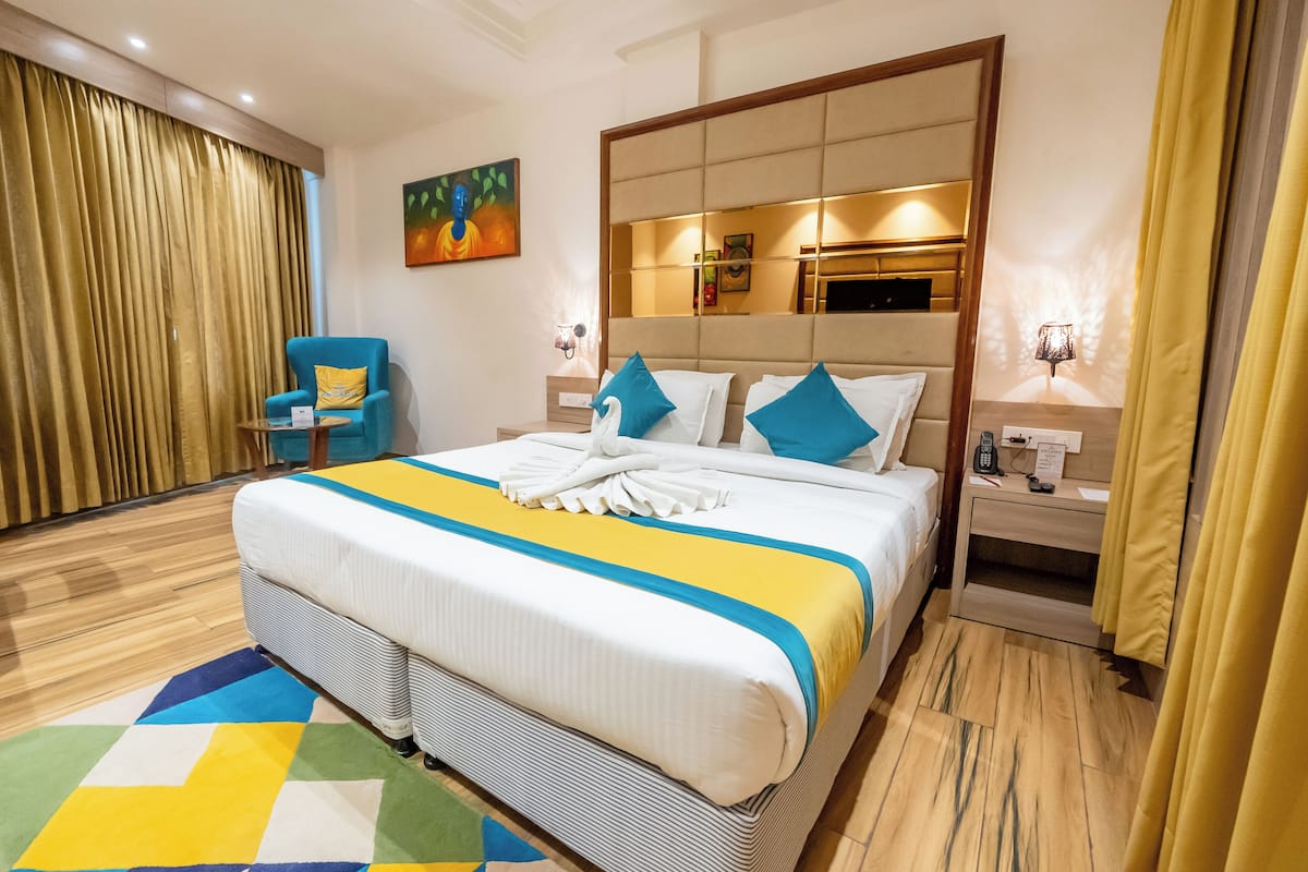 A hotel room with a large bed, a blue armchair, a wooden floor, a painting on the wall, and a mirror above the bed.