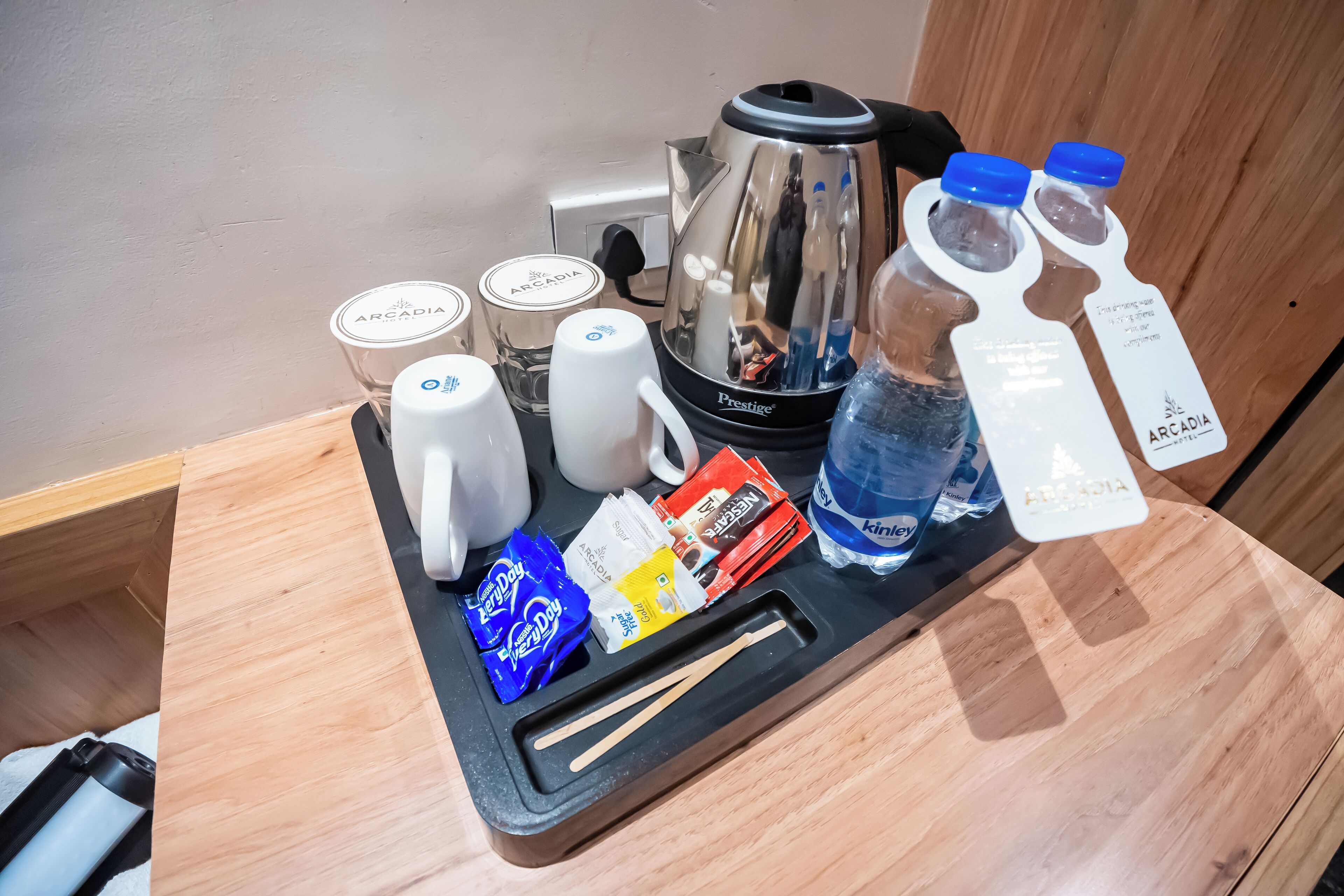 Bandeja de desayuno en una habitación de hotel, con cafetera, tazas, botella de agua y alimentos para el desayuno.