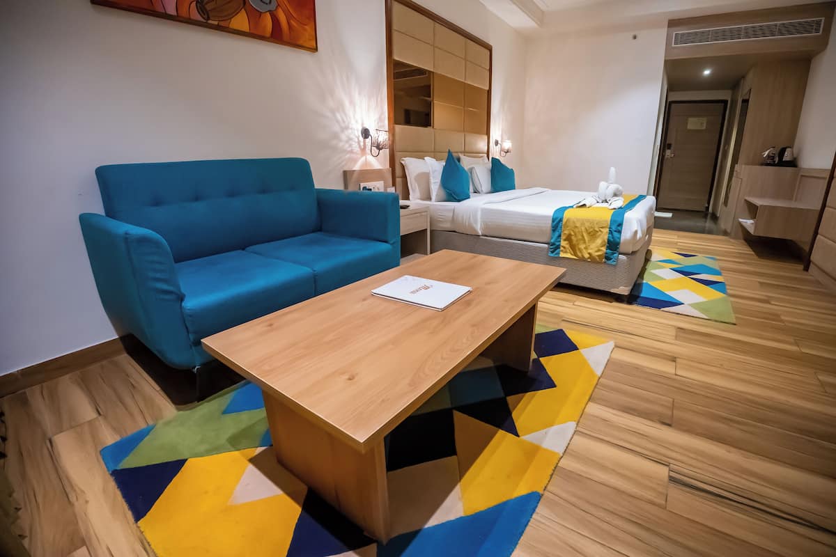A modern hotel room with a blue sofa, a wooden coffee table, a bed with blue and white bedding, and a colorful geometric rug.