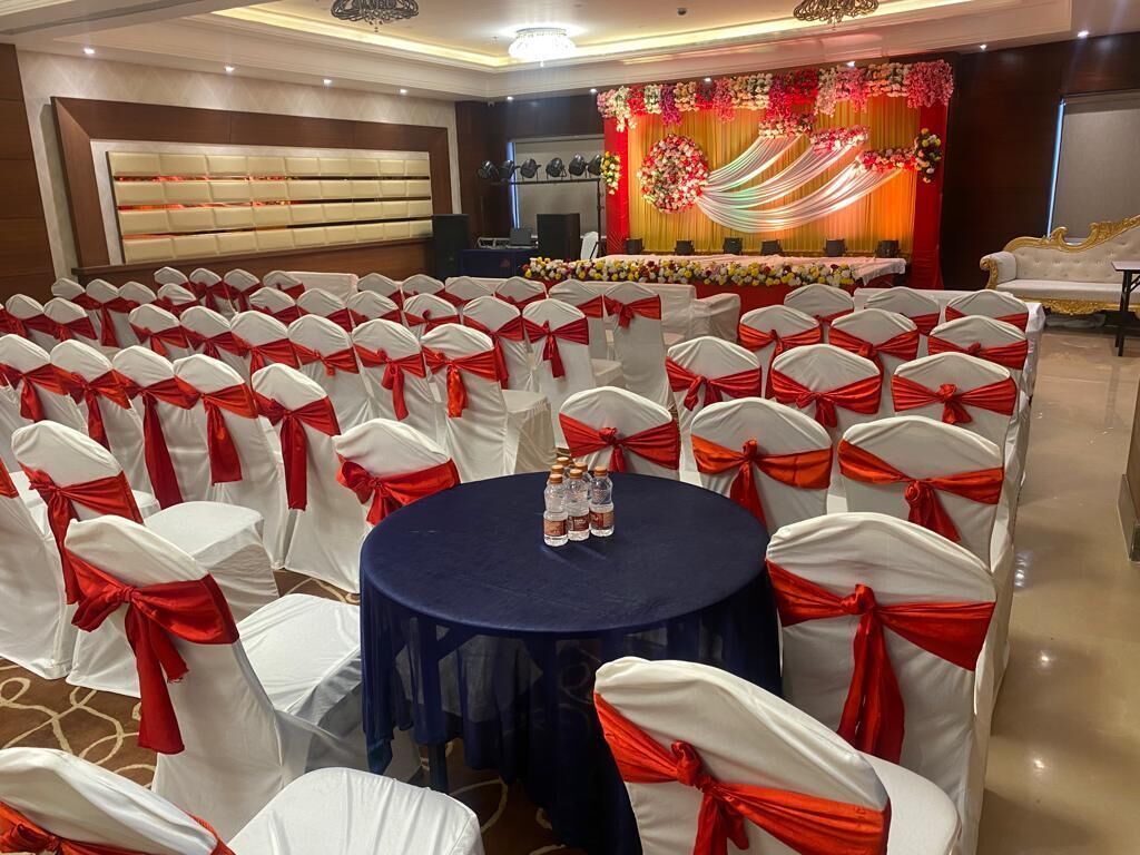 Un salón de banquetes con filas de sillas blancas con cintas rojas, una mesa redonda con botellas y un escenario con decoraciones florales.