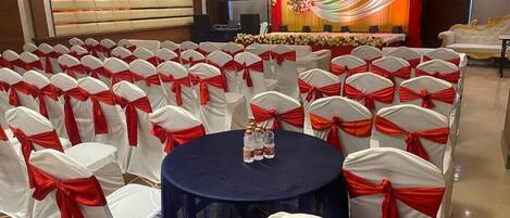 Un salón de banquetes con filas de sillas blancas con cintas rojas, una mesa redonda con botellas y un escenario con decoraciones florales.