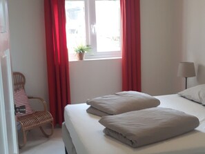 A hotel room with a bed, a chair, a window with red curtains, and a lamp.