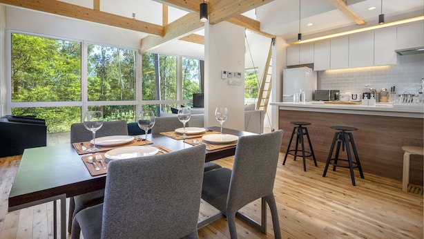 A modern dining area with a wooden table and chairs, large windows offering a view of trees, and a kitchen with white cabinets and a countertop. - Homerunner by H2 Life (Kutchan)