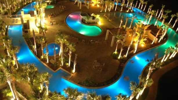Une vue nocturne d’un complexe hôtelier doté de plusieurs piscines, de palmiers et d’espaces bien éclairés.