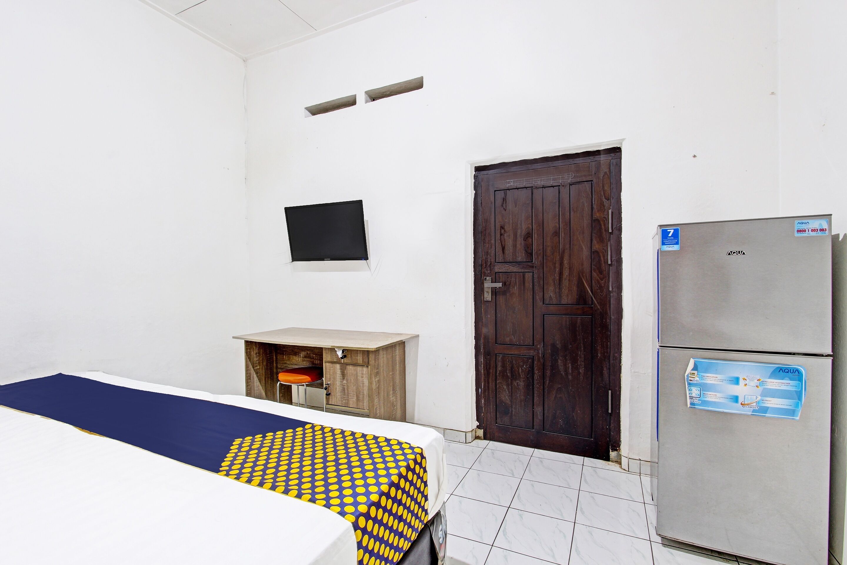 A room with a bed, a wooden cabinet, a dark wooden door, and a refrigerator.