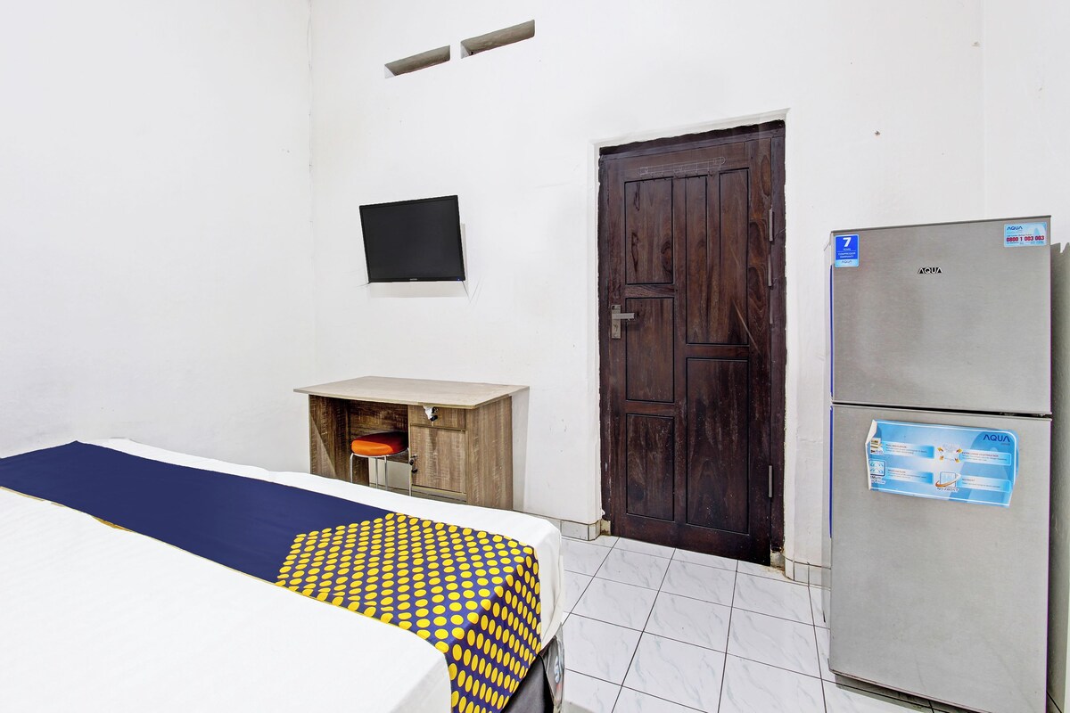 Una habitación con una cama, un armario de madera, una puerta de madera oscura, un televisor montado en la pared y un refrigerador.