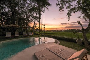 A poolside area with lounge chairs and umbrellas, surrounded by trees and a view of a sunset over a rice field. - Sealuna Villas (Tabanan)