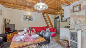 A dining area with a wooden table, chairs, and a red sofa. A stone wall and a stove are visible in the background.