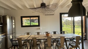 Een eetgedeelte met een grote tafel, stoelen, een hangende lamp, een raam met uitzicht op groen en een keuken met apparatuur.