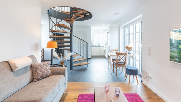 Ein modernes Wohnzimmer mit geschwungener Treppe, einem Sofa, einem Couchtisch und einem Essbereich mit Stühlen und einem Tisch.