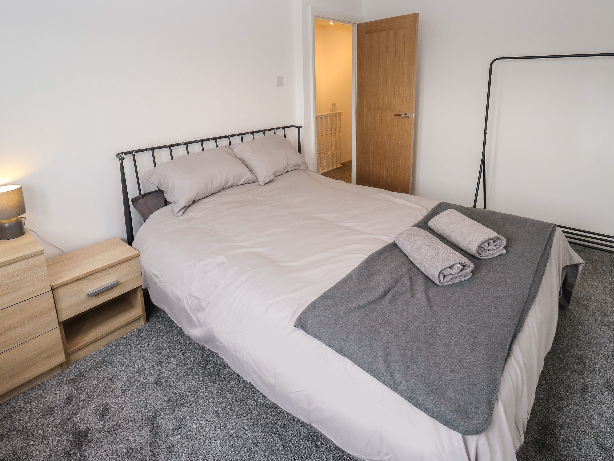 A neatly made bed with a grey blanket and pillows, a bedside table with a lamp, and a wooden wardrobe in the background.