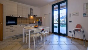 A modern kitchen with white cabinets, a dining table with chairs, a large window with a view of mountains, and a small table with a striped tablecloth. - Appartamento Leone in Vertine (Sulzano)