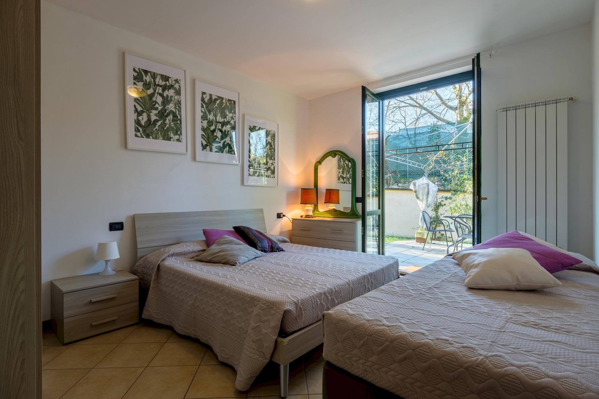 Ein Schlafzimmer mit zwei Betten, einem Nachttisch, einer Kommode und einem Spiegel. An den Wänden hängen Bilderrahmen, und ein Fenster bietet Blick ins Freie.