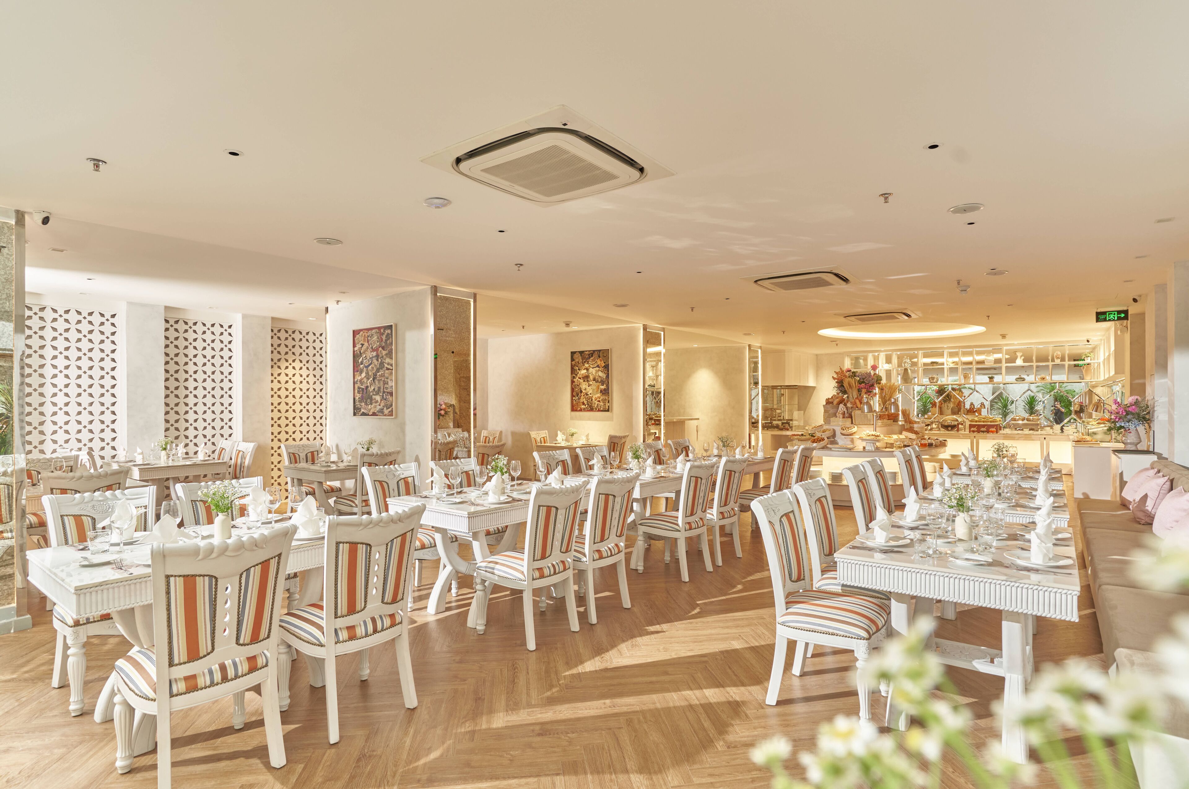 A spacious dining area with multiple tables and chairs, a kitchen in the background, and a decorative wall with geometric patterns.