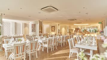 A spacious dining area with multiple tables and chairs, a kitchen in the background, and a decorative wall with geometric patterns.