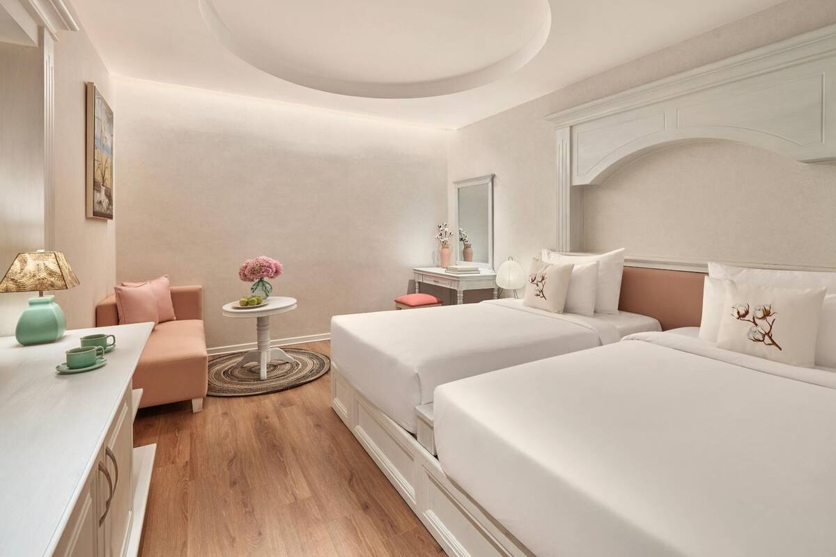 Quarto de hotel com duas camas, uma secretária com luminária, uma mesinha com um vaso e uma cadeira.