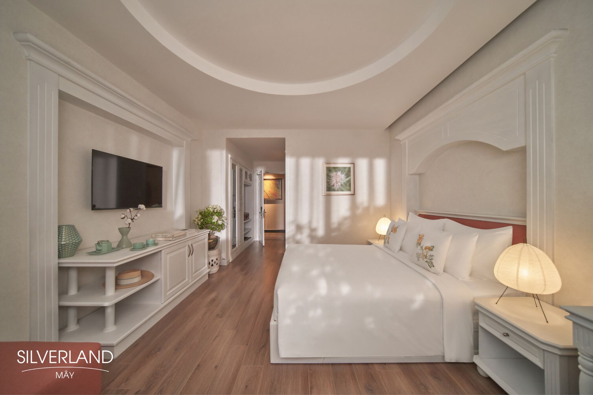 A hotel room with a large bed, a TV mounted on the wall, a bedside table with a lamp, and a view of the hallway.