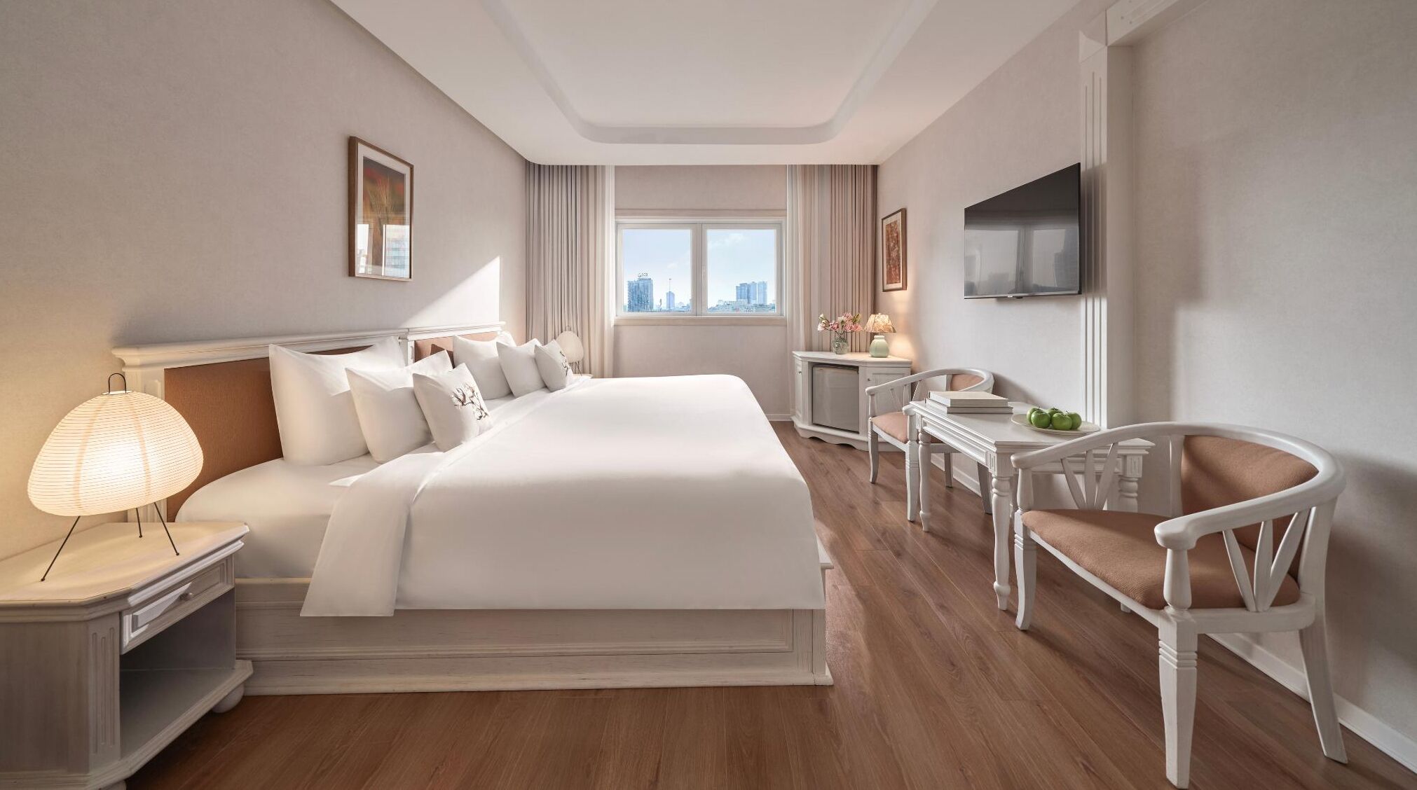 A hotel room with a large bed, a desk, a chair, a TV, and a window with a city view.