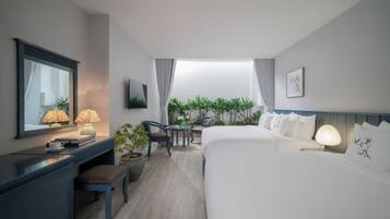 A modern hotel room with a large bed, a desk with a lamp, a chair, a small table, and a view of greenery through the window.