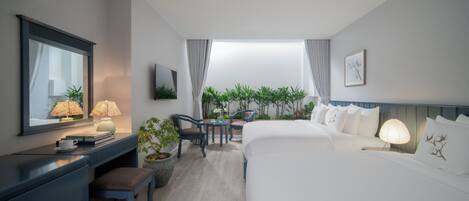A modern hotel room with a large bed, a desk with a lamp, a chair, a small table, and a view of greenery through the window.